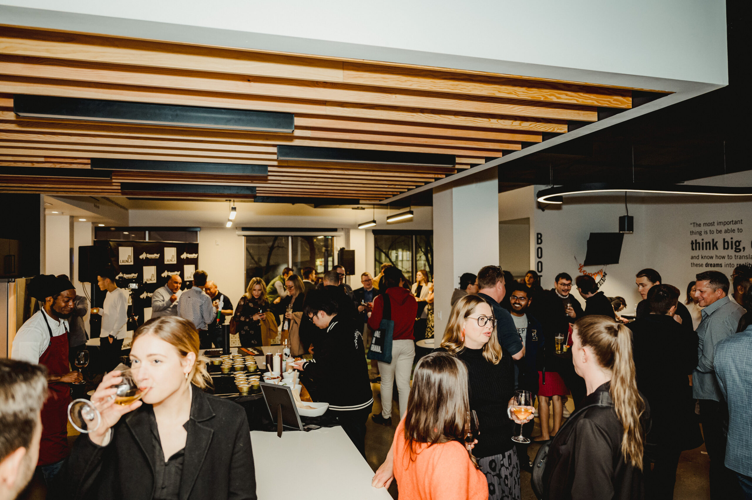 Over 80 people in the Winnipeg business and tech community celebrate the Grand Opening of Launch Downtown (185 Carlton St). There is a banner in the back-left, a classy interior, and people enjoying wine, cocktails, food and conversation in the space.